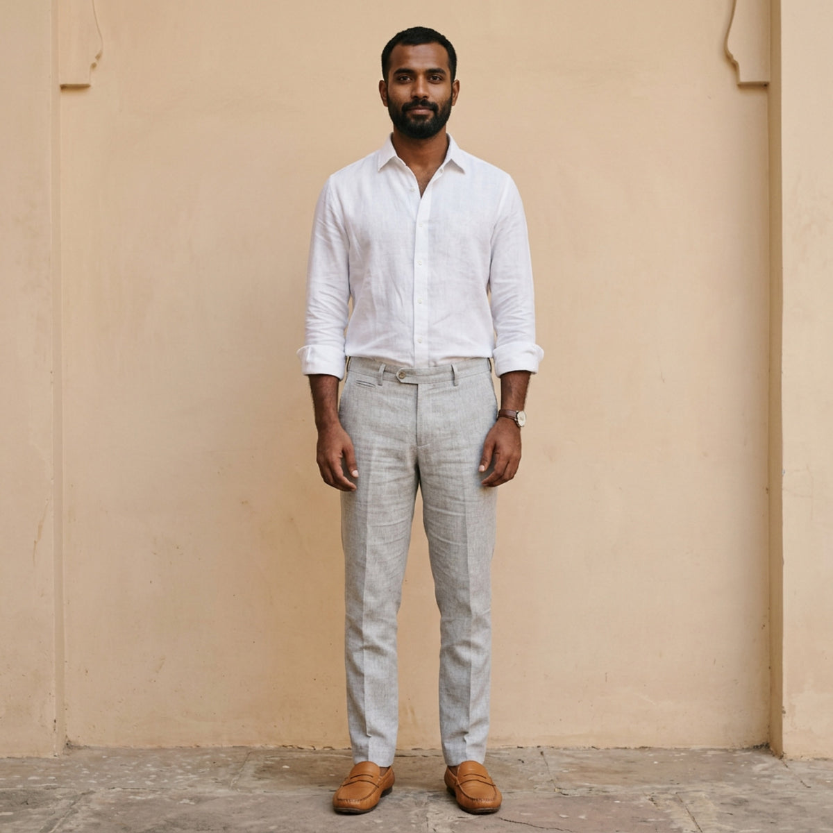 Man in a white shirt and light grey trousers standing against a plain warm wall